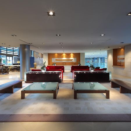 فندق يوروستارز غراند مارينا جي إل برشلونة المظهر الخارجي الصورة The photo shows a modern and spacious interior, likely of a lobby or waiting area. It features several red sofas arranged in a conversational layout, complemented by glass-top coffee tables. The lighting is bright and there are large windows, allowin