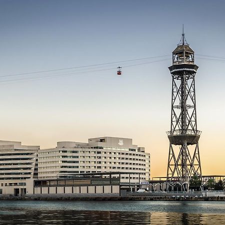 فندق يوروستارز غراند مارينا جي إل برشلونة المظهر الخارجي الصورة The photo shows a modern building alongside a waterfront, with a distinctive tower nearby. The tower appears to be part of a cable car system, featuring a conical roof and a series of cables extending from it. The scene is set against a colorful sky,