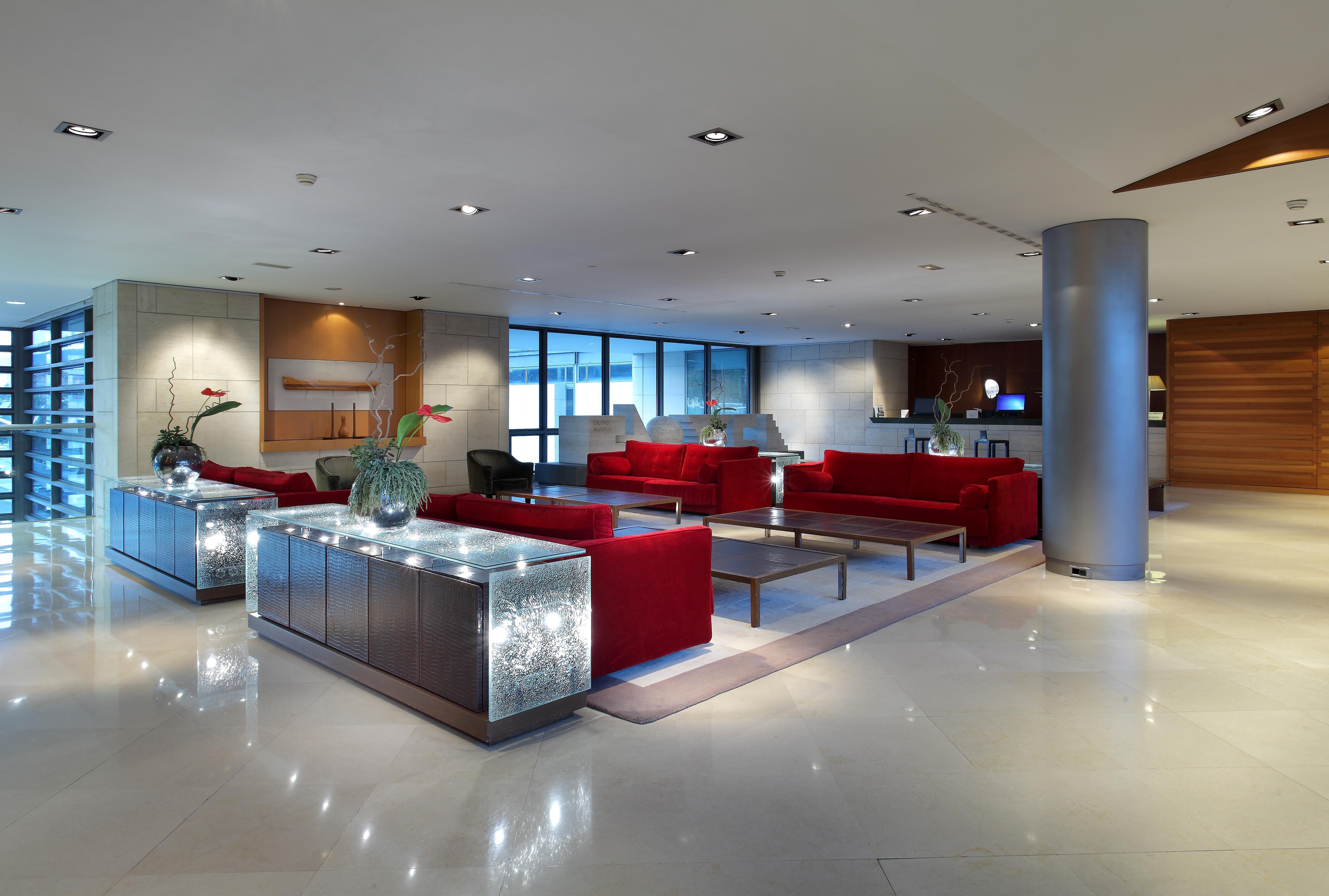 فندق يوروستارز غراند مارينا جي إل برشلونة المظهر الخارجي الصورة The photo depicts a modern lobby area, featuring stylish furnishings. There are several red sofas arranged around coffee tables, creating a comfortable seating area. The flooring looks polished and smooth, with a neutral color that complements the fu