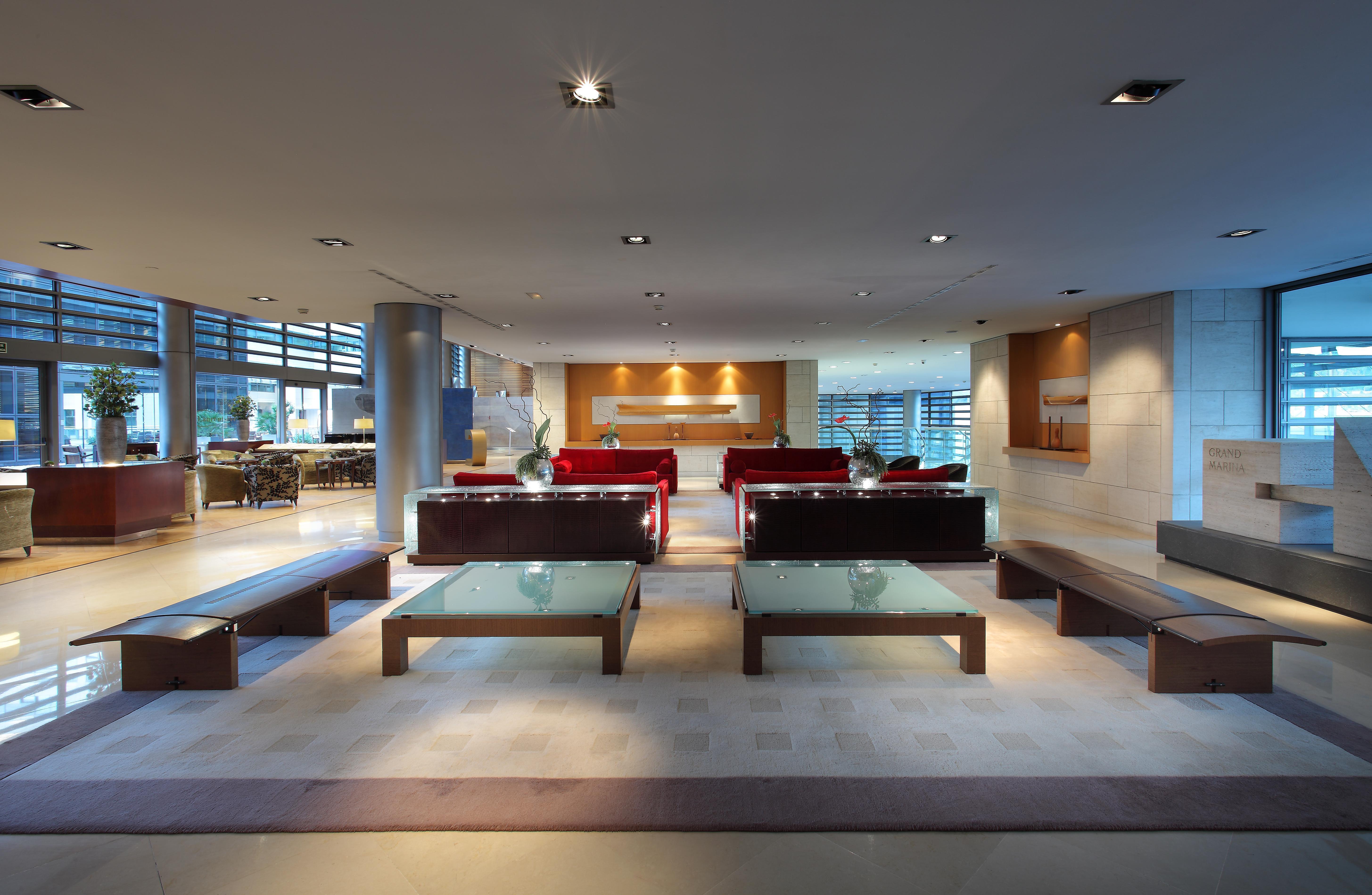 فندق يوروستارز غراند مارينا جي إل برشلونة المظهر الخارجي الصورة The photo shows a modern and spacious interior, likely of a lobby or waiting area. It features several red sofas arranged in a conversational layout, complemented by glass-top coffee tables. The lighting is bright and there are large windows, allowin