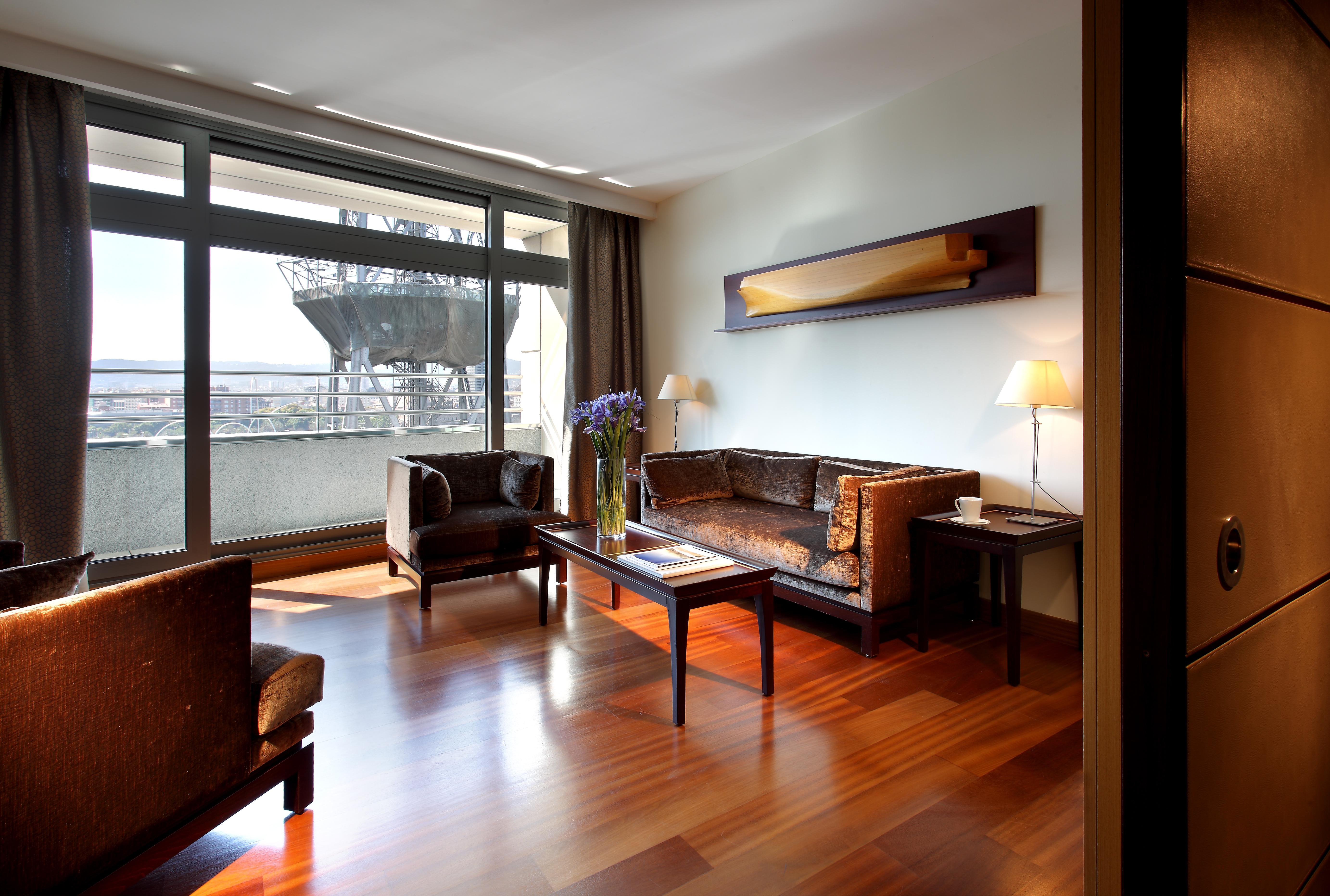 فندق يوروستارز غراند مارينا جي إل برشلونة المظهر الخارجي الصورة The photo depicts a modern living room setting. It features a spacious area with large windows allowing natural light to flood in. The room is furnished with two dark chairs and a brown couch, all arranged around a wooden coffee table. A decorative l