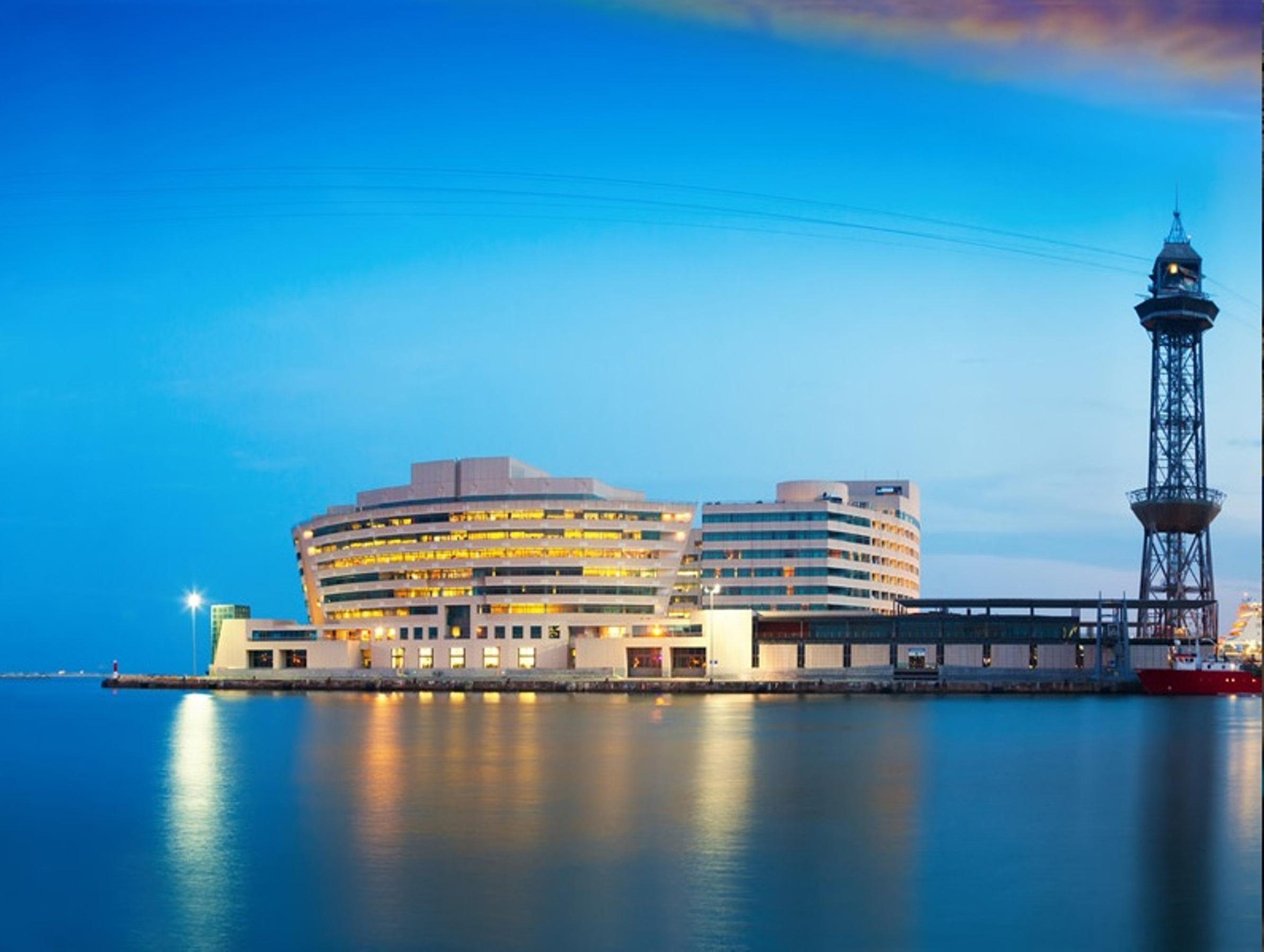 فندق يوروستارز غراند مارينا جي إل برشلونة المظهر الخارجي الصورة The photo shows a modern building situated near the water, likely a harbor or port area. The structure features a sleek, curved design with multiple levels and large windows, reflecting an urban architectural style. Next to the building, there is a t
