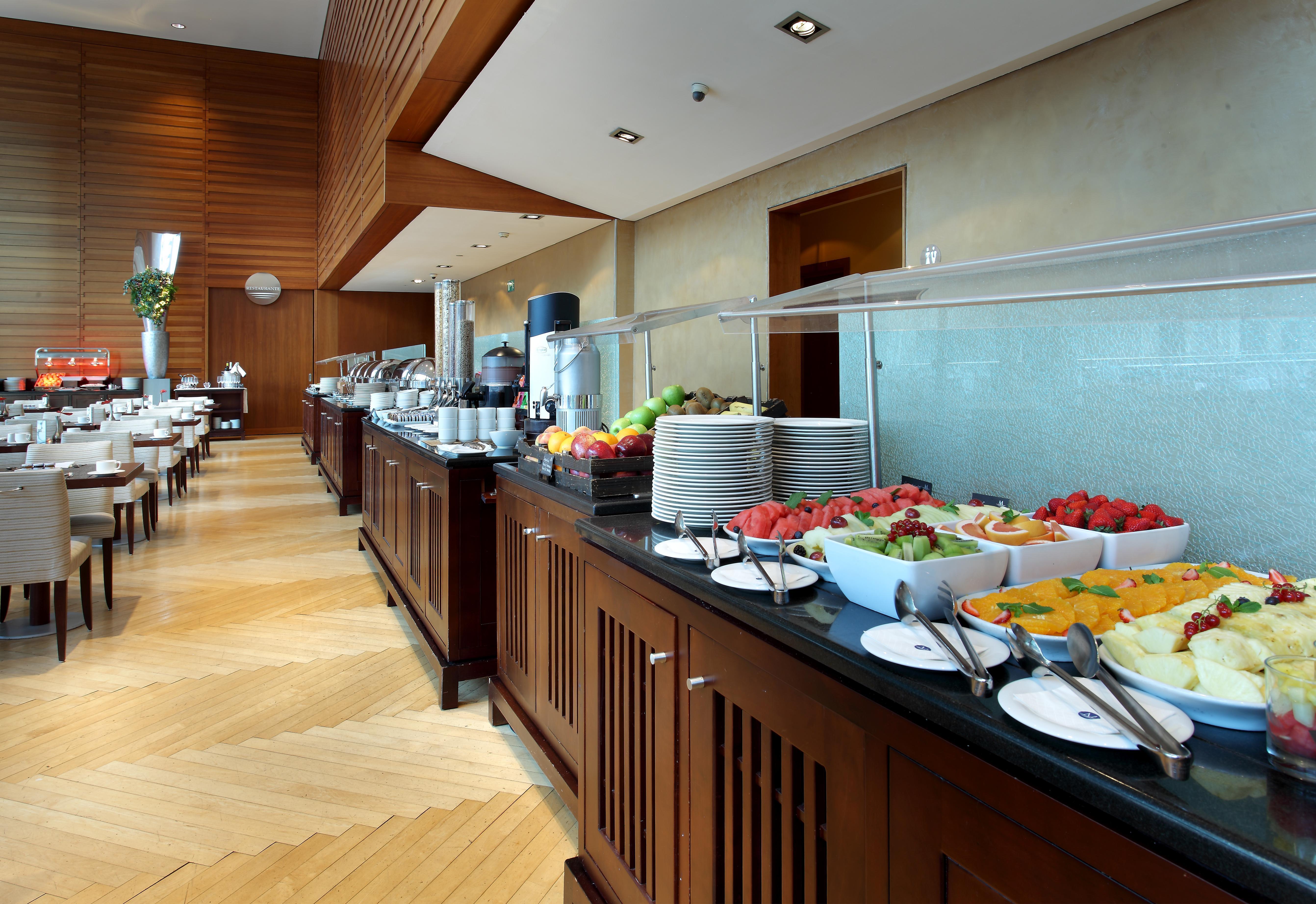 فندق يوروستارز غراند مارينا جي إل برشلونة المظهر الخارجي الصورة The photo shows a well-organized buffet setup in a modern dining area. There is a long counter made of wood, displaying a variety of food items. Several bowls filled with fresh fruits, such as melons and strawberries, are prominently placed. In the b