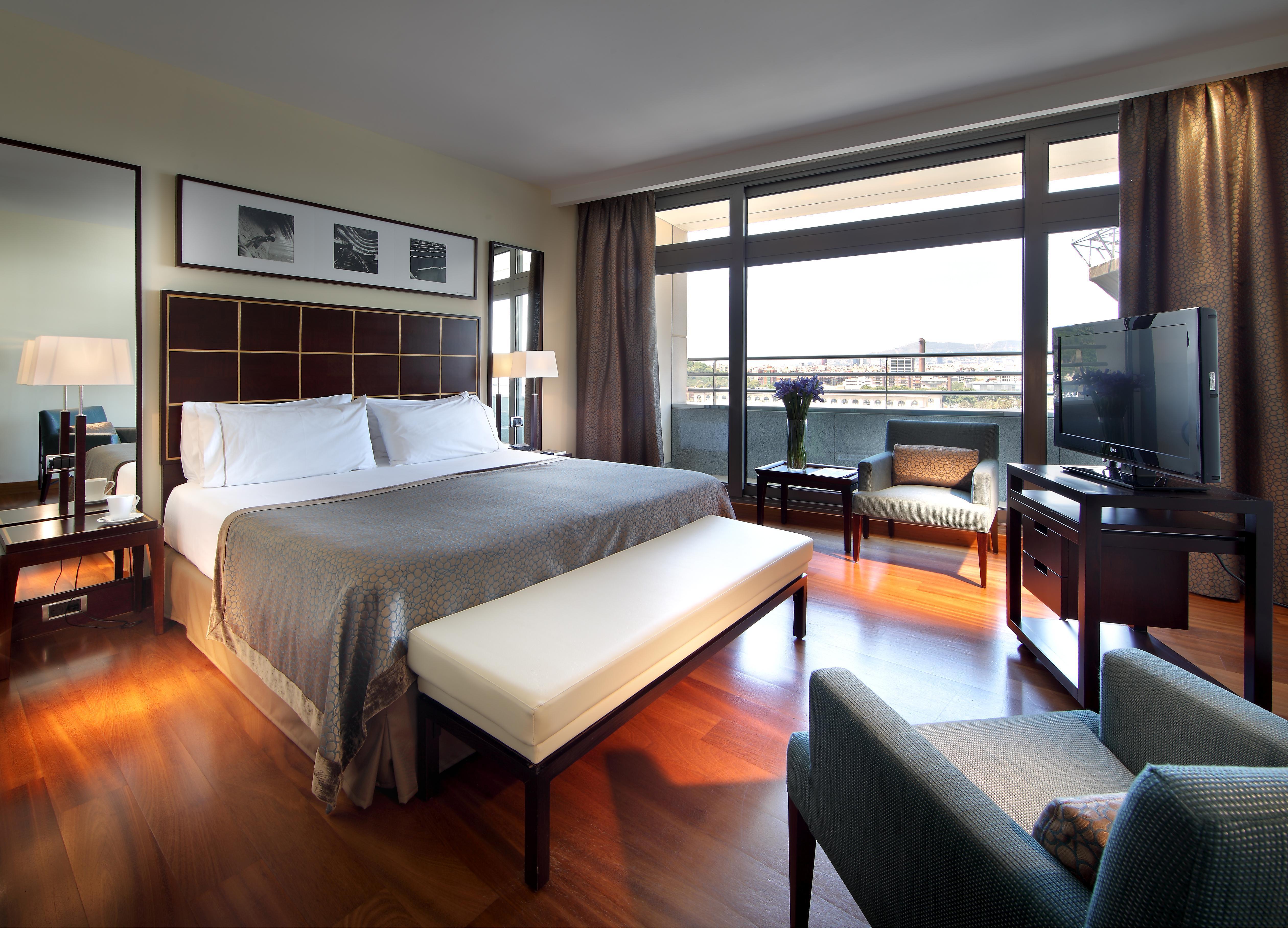 فندق يوروستارز غراند مارينا جي إل برشلونة المظهر الخارجي الصورة The photo shows a cozy hotel room featuring a large bed with white linens and a gray bedspread. There is a light-colored bench at the foot of the bed. A small seating area includes a comfortable chair and a lamp, along with a wooden side table. The r