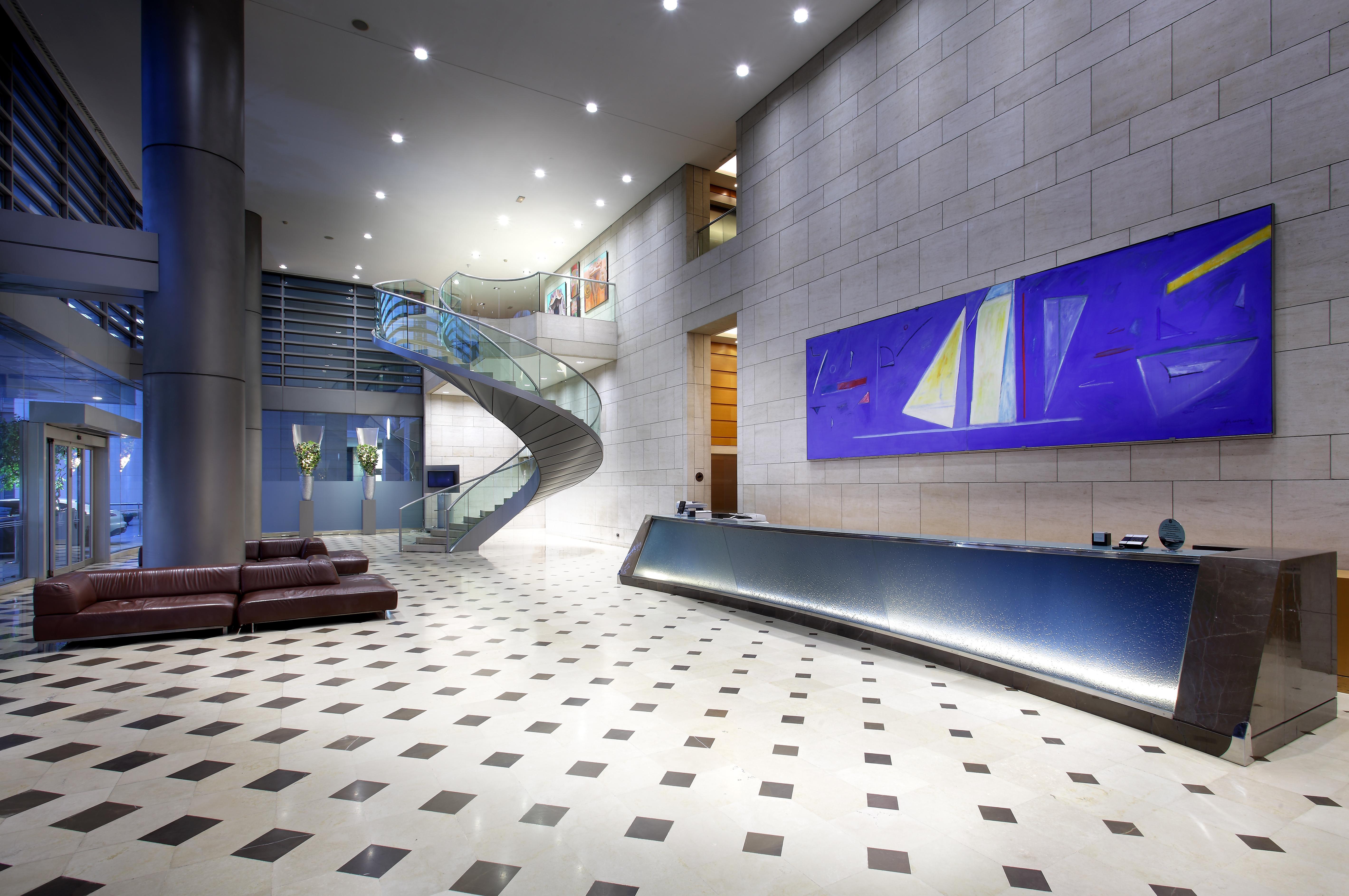فندق يوروستارز غراند مارينا جي إل برشلونة المظهر الخارجي الصورة The photo shows a modern lobby or reception area. There is a sleek reception desk along one wall, featuring a polished surface. To the left, a spacious staircase with a glass guardrail leading to an upper level can be seen, framed by walls decorated 
