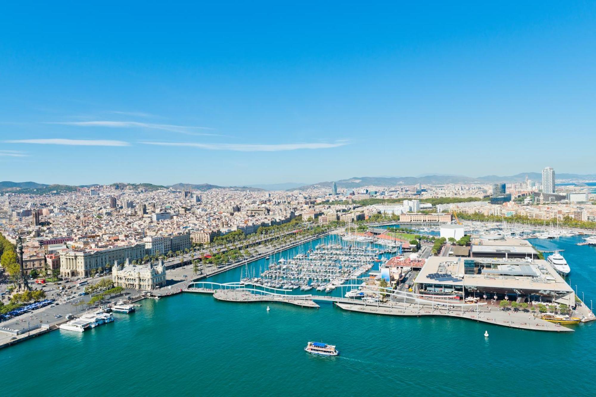 فندق يوروستارز غراند مارينا جي إل برشلونة المظهر الخارجي الصورة The photo depicts a coastal city view, likely of a vibrant harbor or marina. In the foreground, there is a clear blue body of water with a few small boats. The harbor features numerous yachts and boats docked in the marina. 

Surrounding the water ar