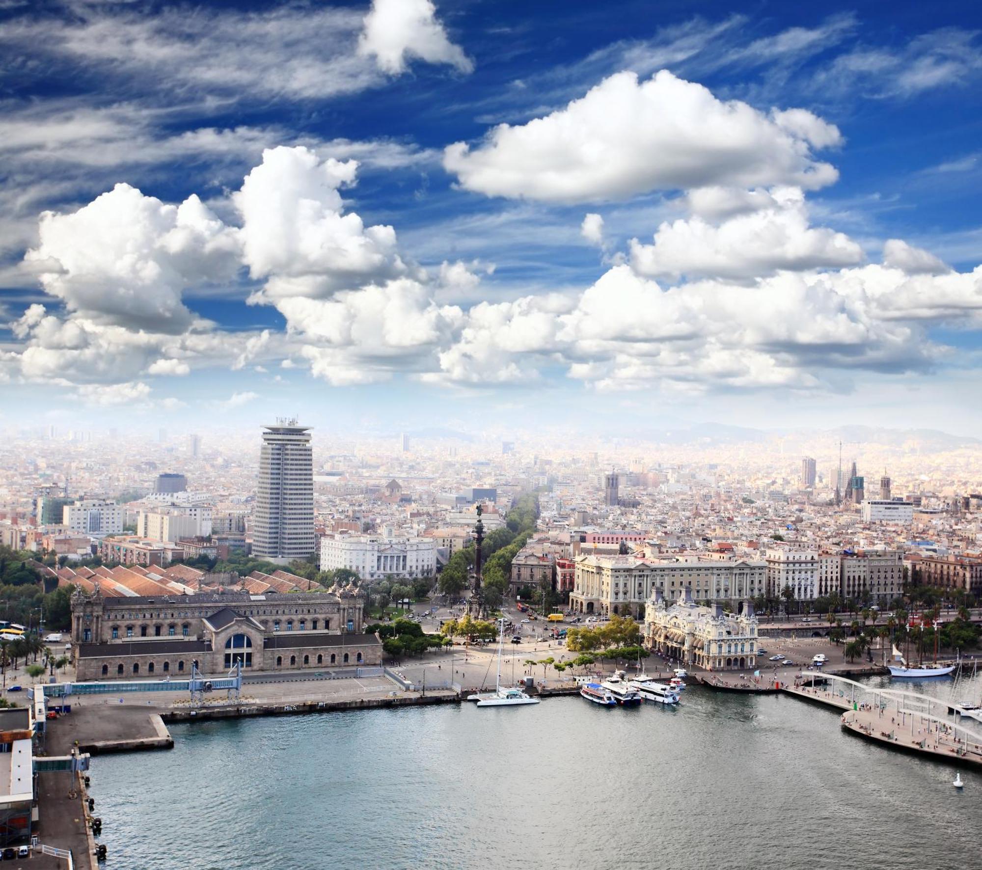 فندق يوروستارز غراند مارينا جي إل برشلونة المظهر الخارجي الصورة The image shows a panoramic view of a cityscape, likely featuring a coastal area with a harbor. In the foreground, there are buildings near the water, including a large structure that might be a historic or notable building. The scene extends into th