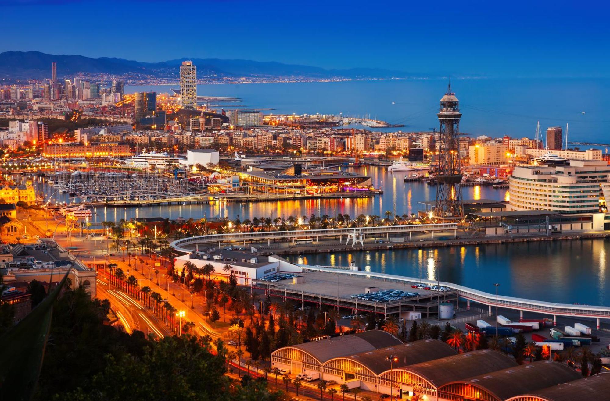 فندق يوروستارز غراند مارينا جي إل برشلونة المظهر الخارجي الصورة The photo depicts a scenic view of a coastal city at dusk. In the foreground, there are illuminated buildings and a marina filled with boats. The waterfront features a curved walkway and a dock area. In the background, you can see a skyline with tall