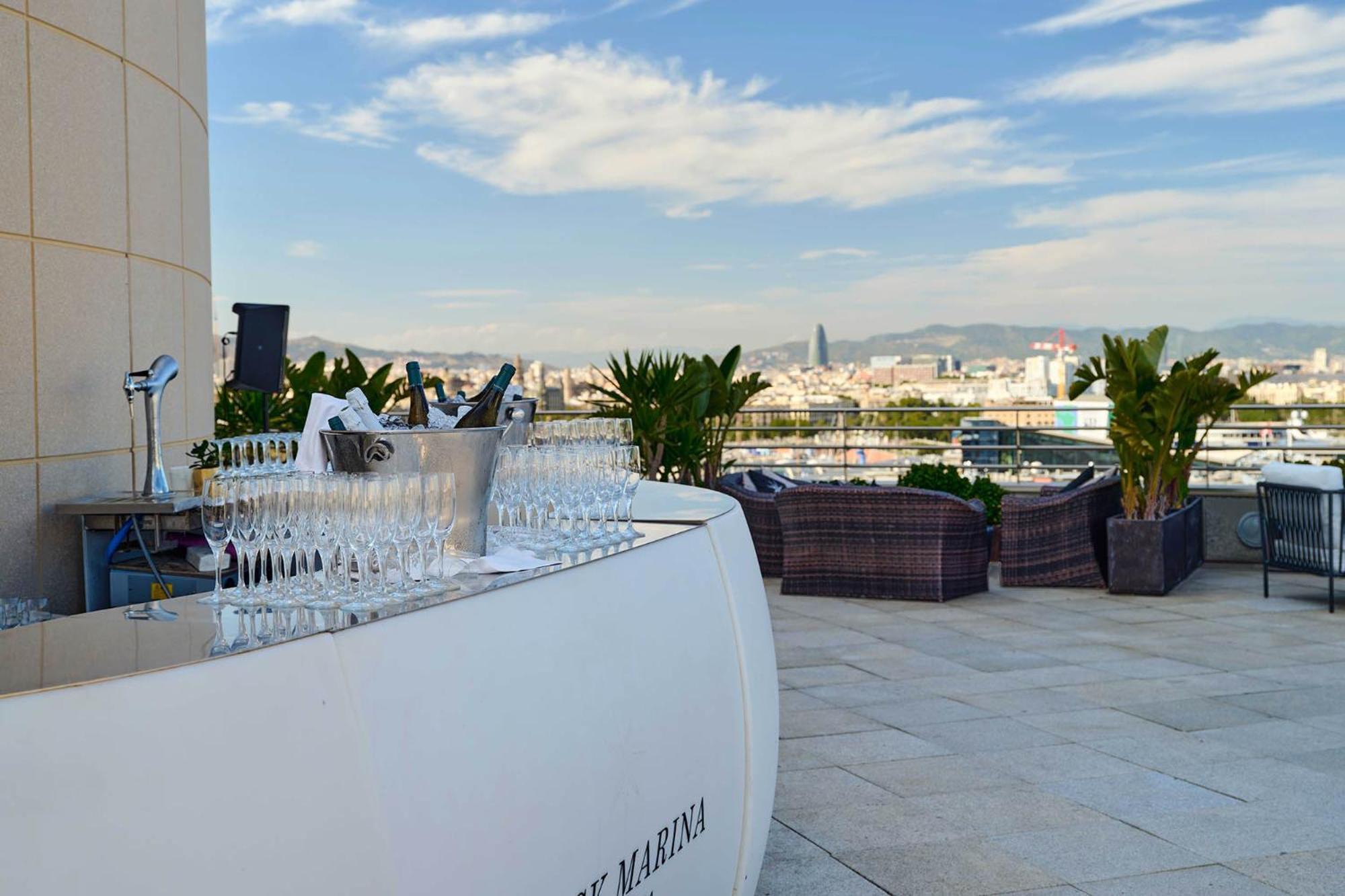فندق يوروستارز غراند مارينا جي إل برشلونة المظهر الخارجي الصورة The photo shows a stylish rooftop bar or terrace area with a modern, curved white bar counter. In front of the bar, there is a row of sparkling glasses lined up for drinks. The background features a beautiful skyline and distant mountains, with a cle