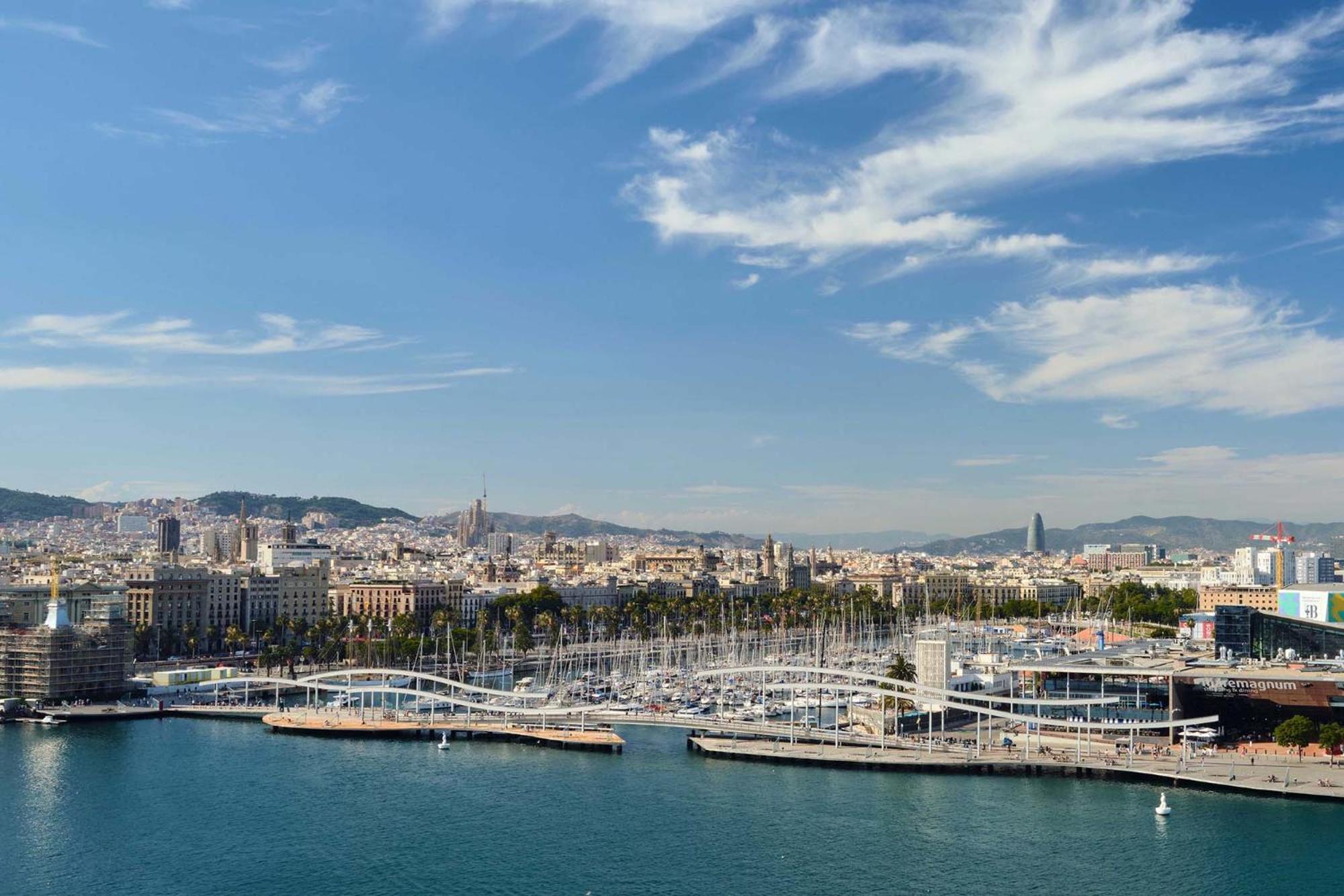 فندق يوروستارز غراند مارينا جي إل برشلونة المظهر الخارجي الصورة The photo shows a scenic view of a coastal city, likely Barcelona, with a harbor area prominent in the foreground. There are several boats moored along the docks, and a distinctive bridge or walkway structure can be seen connecting different parts of