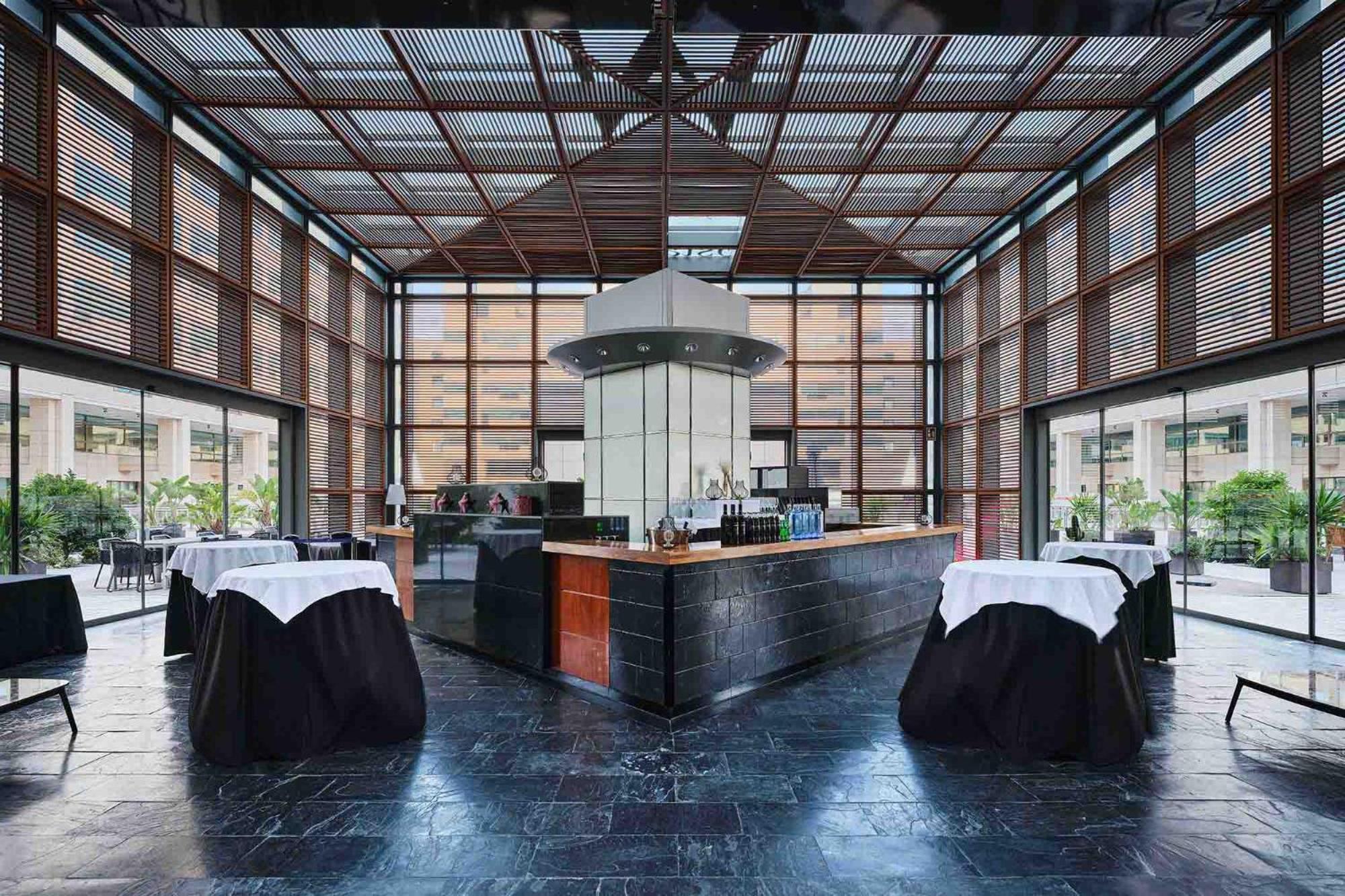 فندق يوروستارز غراند مارينا جي إل برشلونة المظهر الخارجي الصورة The photo shows the interior of a modern restaurant featuring a sleek bar area. The bar is centrally located and has a metallic structure with a contemporary design. Surrounding the bar are several tables covered with dark cloths and white table runn
