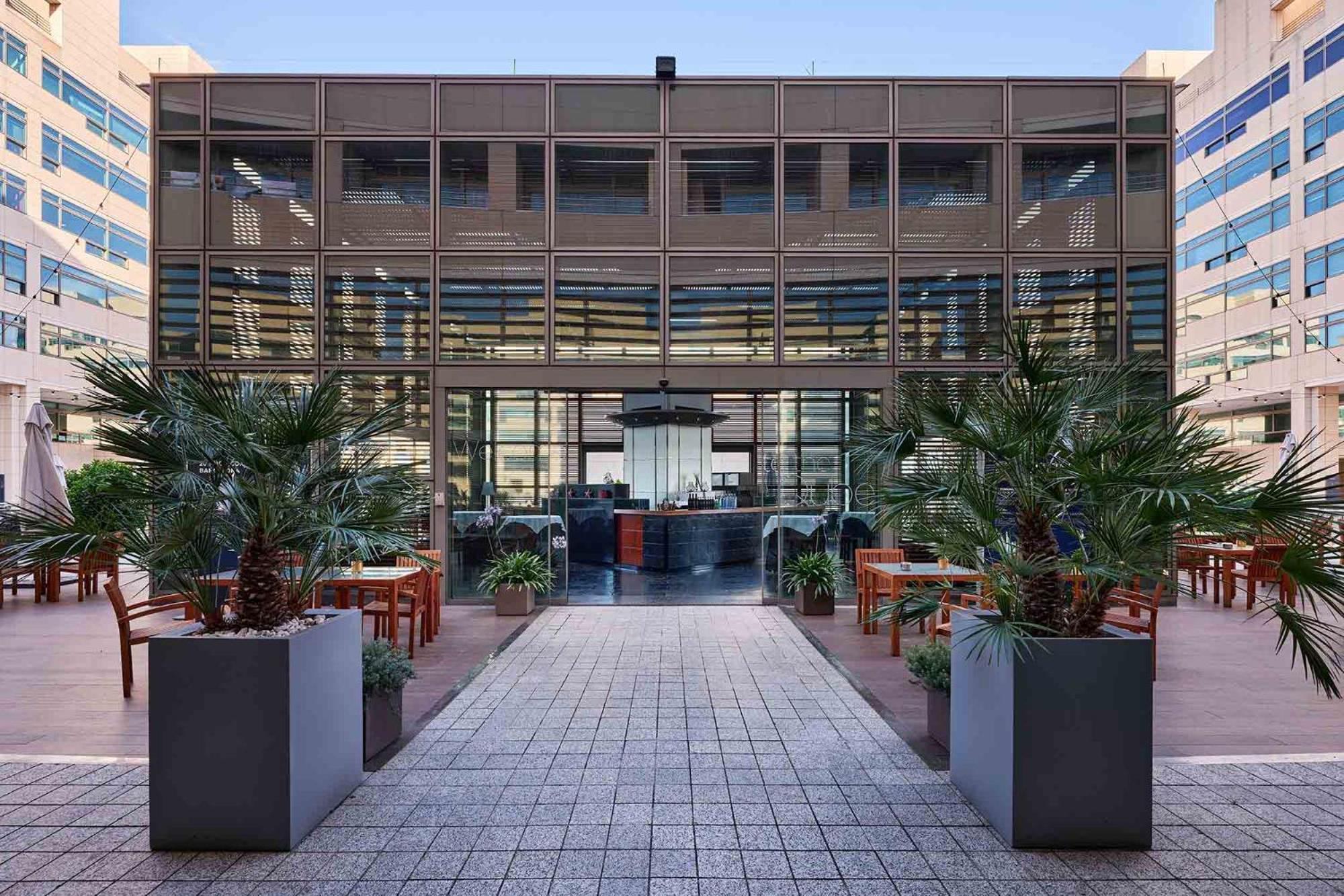 فندق يوروستارز غراند مارينا جي إل برشلونة المظهر الخارجي الصورة The photo depicts a modern building with a glass facade, featuring a central entrance framed by large planters with tropical plants. The entrance leads to an interior space that appears to be a restaurant or café, with a stylish counter and seating v