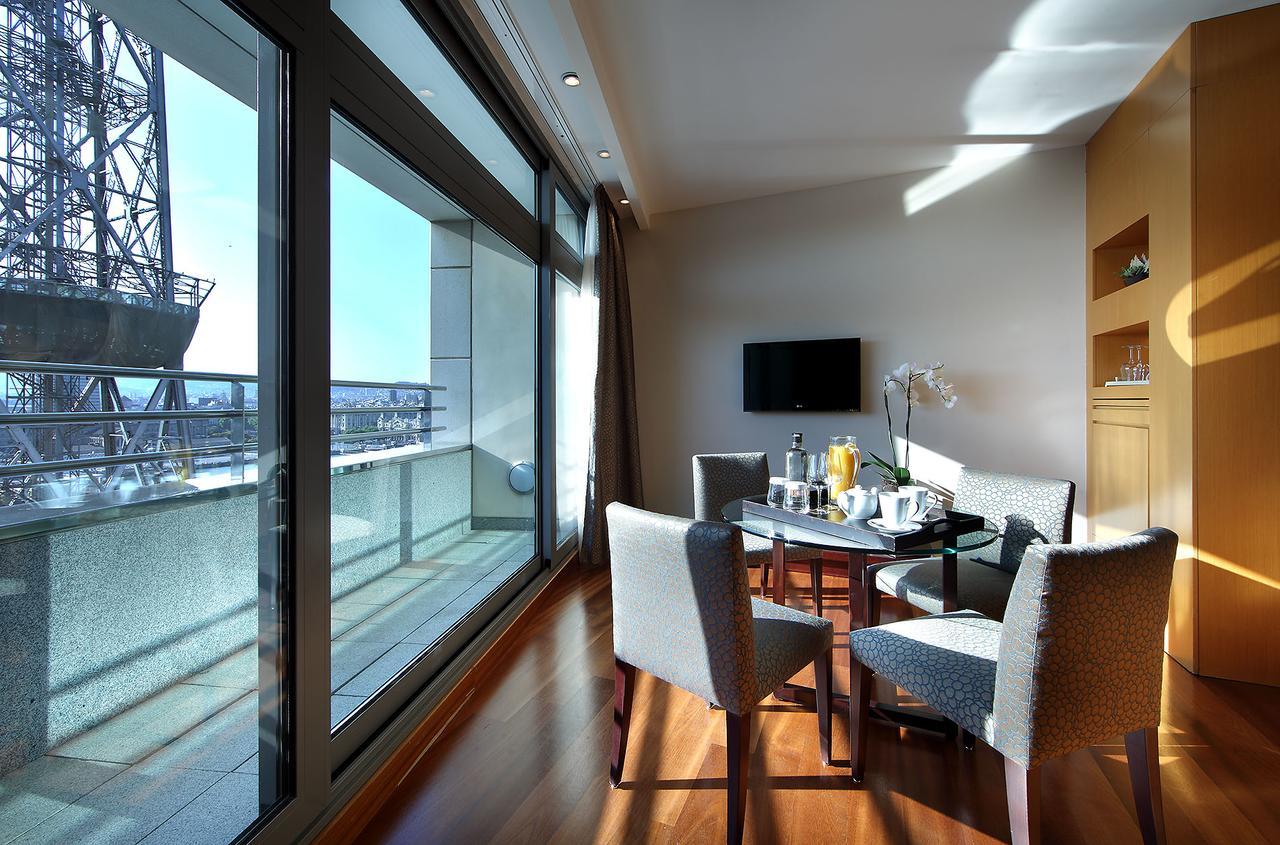 فندق يوروستارز غراند مارينا جي إل برشلونة المظهر الخارجي الصورة The photo shows a modern and bright room that features large windows with panoramic views. Inside, there is a circular dining table surrounded by upholstered chairs. On the table, there are items that suggest a meal or refreshments, such as drinks an