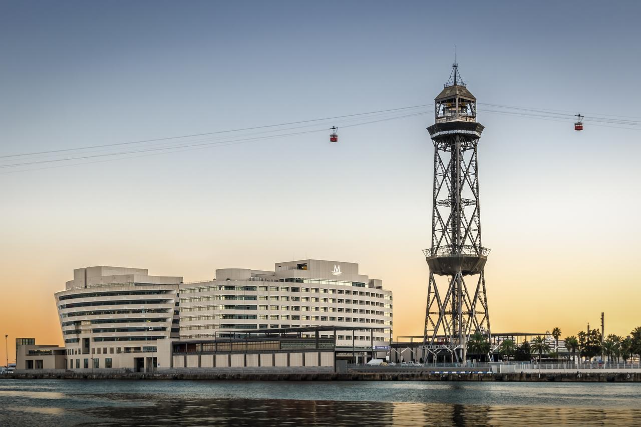 فندق يوروستارز غراند مارينا جي إل برشلونة المظهر الخارجي الصورة The photo shows a modern building alongside a waterfront, with a distinctive tower nearby. The tower appears to be part of a cable car system, featuring a conical roof and a series of cables extending from it. The scene is set against a colorful sky,