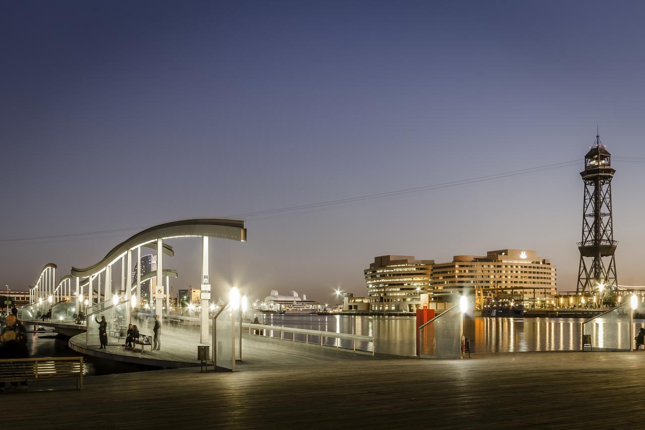 فندق يوروستارز غراند مارينا جي إل برشلونة المظهر الخارجي الصورة The photo depicts a scenic waterfront view during twilight. On the left, there is a modern, curved pedestrian walkway that appears to be well-lit, inviting pedestrians. To the right, a tall structure resembling a lighthouse or observation tower stand
